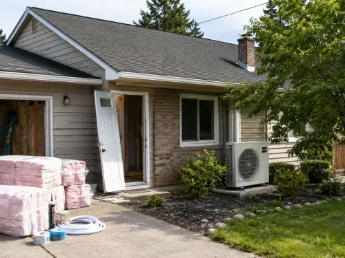 Modest house being upgraded with insulation, air-sealing supplies, heat pump equipment, shade planting, and a solar-ready roof.
