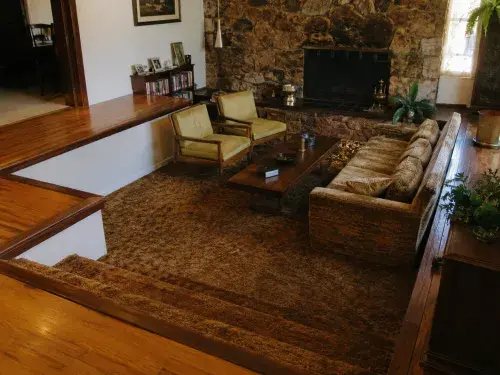 1970s sunken living room with carpeted conversation pit, fireplace, sofa, chairs, and upper floor walkway.
