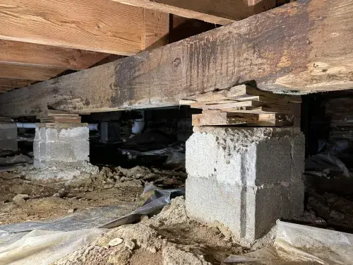 Pier and beam foundation repair inspection inside a crawl space with wood beams, stacked shims, masonry piers, dirt floor, and torn vapor barrier.