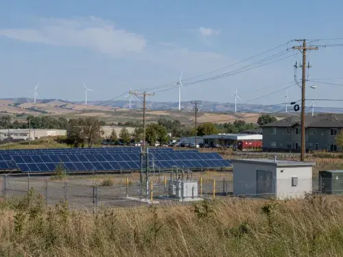 Solar panels, wind turbines, utility lines, service equipment, and nearby buildings showing how renewable energy connects to real infrastructure.