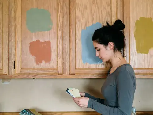 Woman comparing paint colors on oak kitchen cabinets with test swatches.