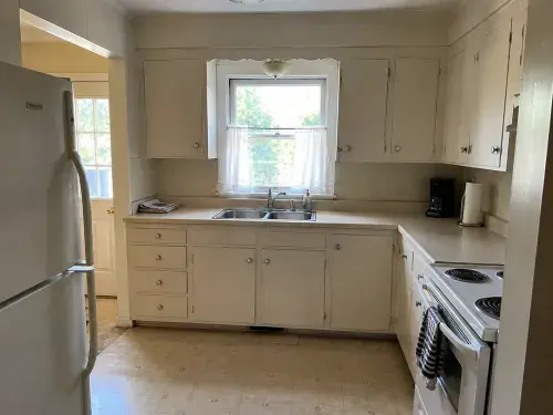 Older small Cape kitchen with sink window, worn cabinets, range, fridge, and tight layout.