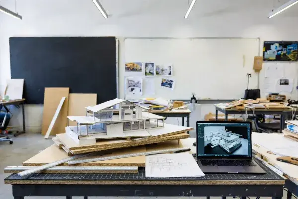 Architecture student studio workspace with physical massing models, drawings, and laptop.