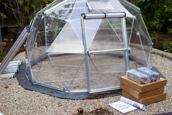 Greenhouse dome kit with visible frame, polycarbonate panels, door, and boxed components on a gravel base.