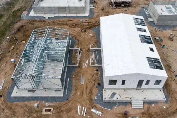 Prefab metal building shell beside a finished wall-panel structure on a construction site.