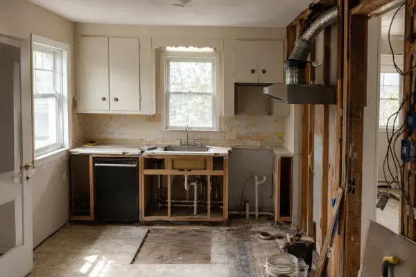 1950s kitchen remodel in progress with exposed wall framing, plumbing, ductwork, old cabinets, floor layers, and unfinished repair areas.