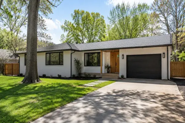 Updated 1960s ranch house exterior with white brick, dark trim, wood entry detail, black garage door, mature trees, driveway, and simple front yard landscaping.