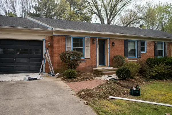 Brick ranch house exterior with taped trim, dark garage door, ladder, tools, and shrubs during an exterior update.