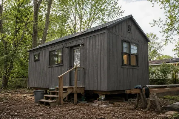 Small dark tiny house in a wooded yard with simple siding, metal roof, temporary supports, entry steps, and construction materials nearby.