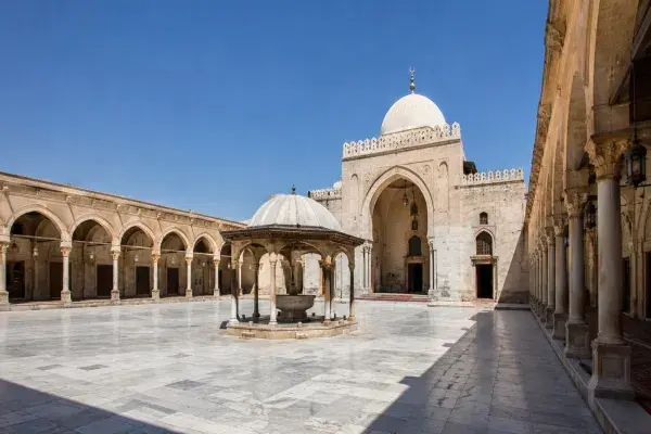 Open mosque courtyard with shaded arcades, central fountain, stone paving, and prayer hall approach showing how a sahn organizes light and shade.