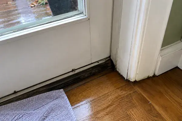 Wet interior floor at the bottom of an exterior door threshold, with water staining near the sill, worn trim, and a towel placed nearby after rain.