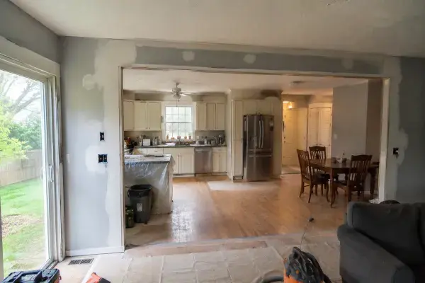 Older ranch kitchen opened to a dining and living area after wall removal, with patched drywall, floor changes, tools, and unfinished repairs.