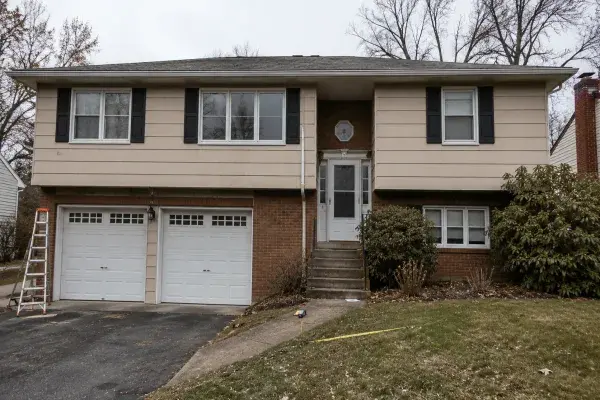 Older raised ranch exterior with dominant garage doors, split entry stairs, small lower windows, dated siding, and overgrown shrubs.