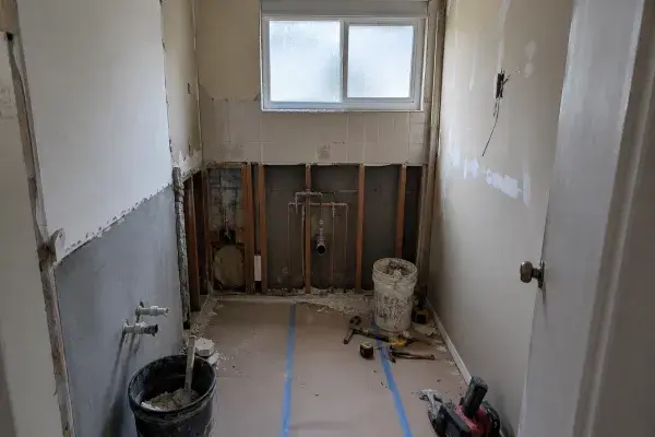 Older ranch bathroom mid-remodel with exposed wall framing, rough plumbing, old tile, patched drywall, tools, and protected flooring.