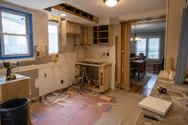 Ranch kitchen mid-remodel with opened soffit, partial cabinet removal, exposed wall patches, protected floor, taped window, tools, and dining room beyond.