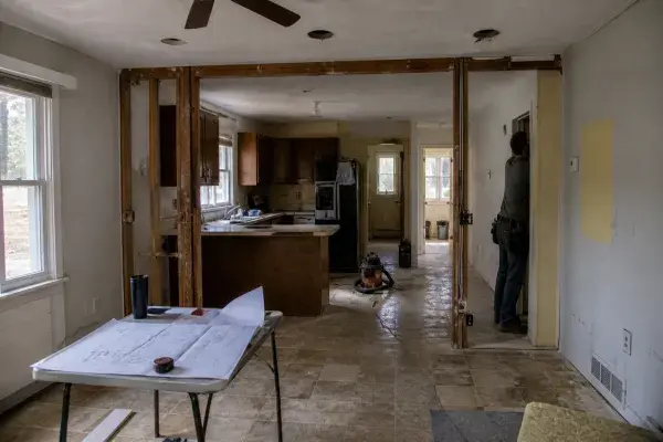 Small ranch house interior remodel with partial wall opening, exposed framing, low ceiling, old kitchen, and plans on a table.