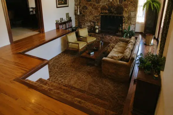 1970s sunken living room with carpeted conversation pit, fireplace, sofa, chairs, and upper floor walkway.