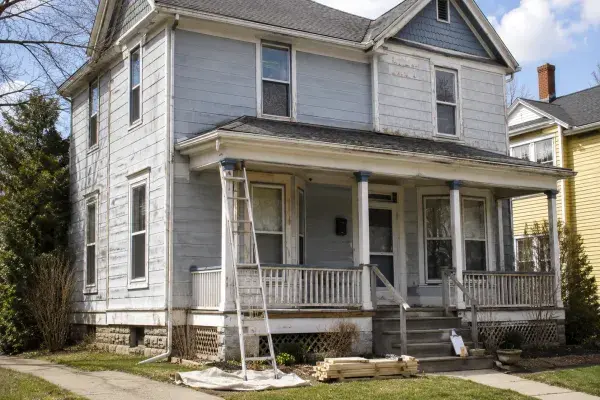 Early-1900s wood house in bright daylight with weathered siding, a covered porch, ladder, lumber and visible exterior repair planning.