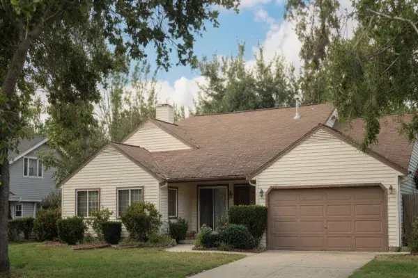 1980s suburban ranch house with cream siding, brown garage door, driveway, lawn, and shrubs.