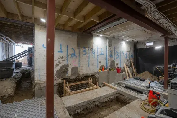 Basement underpinning and lowering project with staged excavation bays, soil removal access, exposed utilities, and new concrete support work.