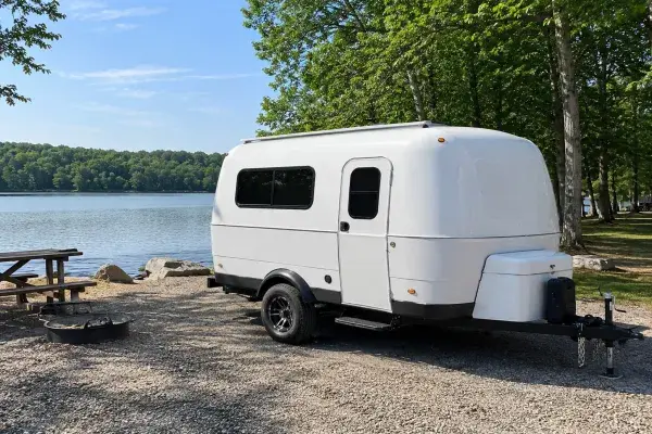 White travel trailer parked beside a lake at a wooded campsite.
